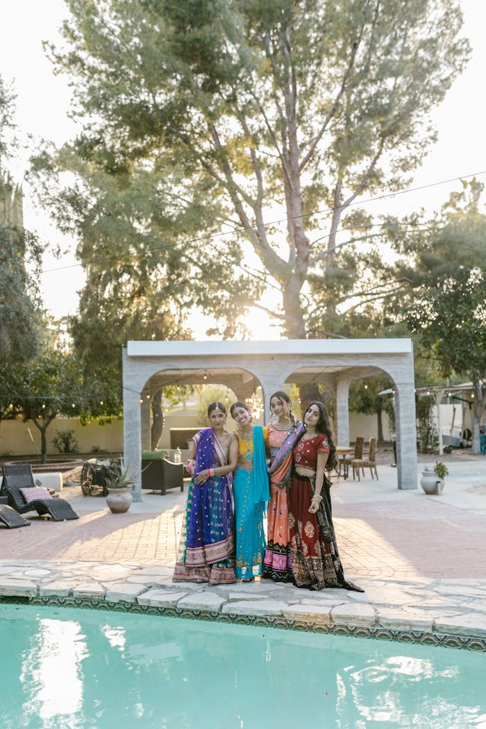 A group of women in colorful traditional sarees posing poolside at sunset, celebrating together.